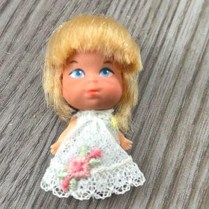 May include: A small doll with blonde hair and blue eyes. The doll wears a white lace dress with a pink floral design. The doll is a vintage toy, likely from the 1960s or 1970s.