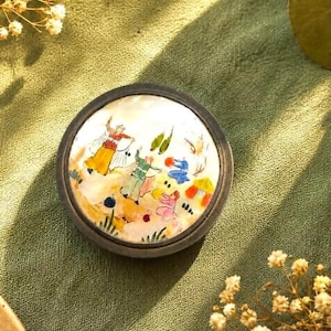 May include: A round, dark-rimmed compact featuring a white, iridescent surface decorated with a colorful, hand-painted scene of several figures in traditional attire engaged in an outdoor activity, set against a light, textured background.