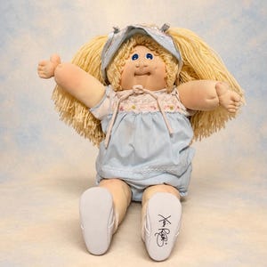 Pode incluir: Uma boneca de pano com cabelo de lã loira, olhos azuis e sardas. A boneca veste um vestido azul claro com corpete de estampa floral, uma touca combinando com fitas e sapatos brancos. O sapato esquerdo apresenta a assinatura Xavier Roberts.