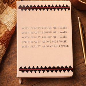 May include: A beige journal with a decorative black and red geometric border. The journal's cover features the text "WITH BEAUTY BEFORE ME I WALK" and other related phrases in a silver font. A gold pen rests nearby.