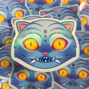 Peut inclure: Autocollant de chat bleu avec de grands yeux orange et des dents pointues. Le chat a un nez rose et des moustaches. L'autocollant a un effet holographique, avec une finition brillante et irisée. Le pelage du chat a un motif rayé.