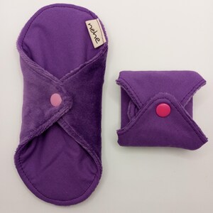 Puede incluir: Compresa menstrual de tela reutilizable de color morado con un botón rosa y una etiqueta que dice "nehe". La compresa está doblada por la mitad y está junto a una compresa morada más pequeña que también está doblada por la mitad.