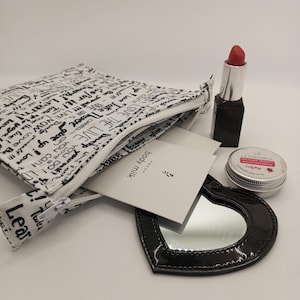 Puede incluir: Una bolsa blanca con cremallera con texto negro, una pequeña tarjeta blanca con texto negro, un espejo compacto negro en forma de corazón, un recipiente plateado con una etiqueta roja y blanca, y un lápiz labial rojo en un tubo plateado y negro.