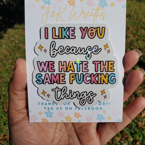 May include: A sticker with the text "I LIKE YOU because WE HATE THE SAME FUCKING things" in a playful font. The sticker is held in a hand, with a floral background and the words "J&K Wreaths" at the top.