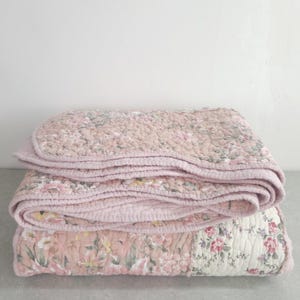 May include: A folded quilted blanket featuring a floral pattern with shades of dusty pink, cream, and green. The fabric has a visible stitched texture throughout, with a repeating botanical print of flowers and leaves across the entire surface.