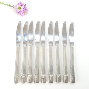 May include: A set of eight silver butter knives with a decorative handle design. The knives are arranged in a row on a white surface, with a sprig of purple flowers in the upper left corner.