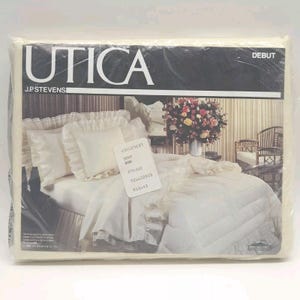 May include: A packaged bedding set featuring the brand name "UTICA" and the word "DEBUT". The set includes a bedspread and pillowcases with ruffled edges, all in a cream color. The packaging is clear, showing the contents.