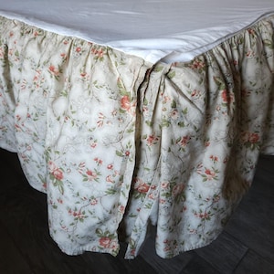 May include: A bed skirt featuring a cream-colored fabric with a repeating floral pattern in shades of pink, green, and brown. The skirt has a gathered, ruffled design that hangs from the edge of a white mattress, creating a decorative border.