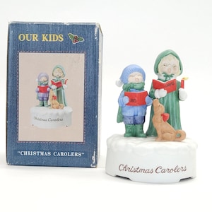 May include: A ceramic Christmas figurine featuring two children in winter attire singing carols from red books, accompanied by a dog. The figurine is on a snowy base with the words "Christmas Carolers". The box reads "Our Kids" and "Christmas Carolers".