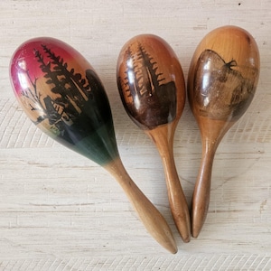 May include: Three wooden maracas with smooth, rounded heads and slender handles. The left maraca features a red and green gradient with a black silhouette of pine trees. The middle maraca shows a dark brown pine tree design, and the right maraca displays a dark brown mountain landscape illustration.