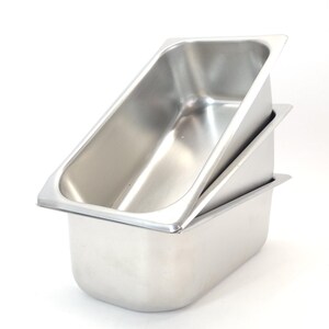 May include: Two stainless steel rectangular food pans stacked on top of each other. The pans have a shiny, reflective surface and are designed with a slightly angled rim. The pans are empty and ready for use.