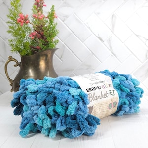 May include: A skein of Bernat Alize Blanket-EZ yarn in shades of blue, with a label visible. The yarn is a loop-style chenille, perfect for creating soft blankets. A vintage brass pitcher with pink flowers is in the background.