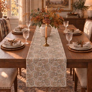 May include: A long rectangular table runner with a muted floral pattern in shades of beige, tan, and soft blue. The runner is centered on a wooden dining table set with white plates, linen napkins, and clear glassware for a formal table setting.