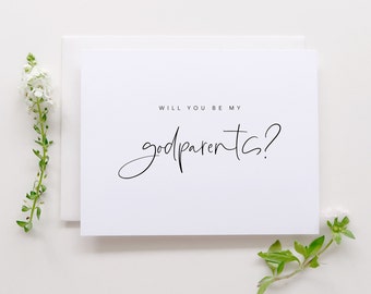 May include: A white card with black text that reads "Will you be my godparents?" The card is on a white background with a small white flower on the right side of the card and a small white flower on the left side of the card.