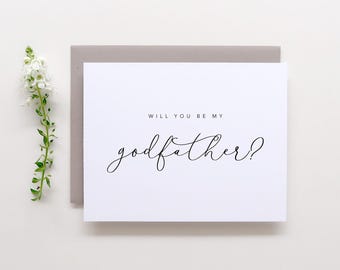 May include: A white greeting card with the black printed text "WILL YOU BE MY godfather?" in a combination of sans-serif and script fonts. The card is placed on top of a gray envelope, accompanied by a single stem of small white flowers.