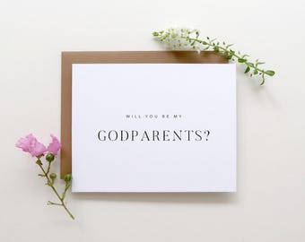 May include: A white rectangular greeting card with the black text WILL YOU BE MY GODPARENTS? centered on the front. The card is placed against a brown envelope, accompanied by a small sprig of white flowers and a stem with pink flowers.