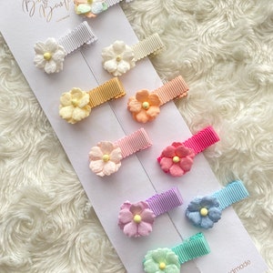 May include: A set of ten pastel-coloured hair clips featuring paper flower embellishments. The flowers are in various shades of pink, blue, yellow, and green. The clips are arranged on a white background.