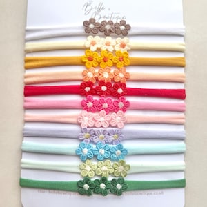 May include: A selection of baby headbands in a variety of colours, each featuring a small, embroidered flower. The headbands are presented on a white card, displaying a spectrum of shades from muted to bright. The brand name "Belle Boutique" is visible.