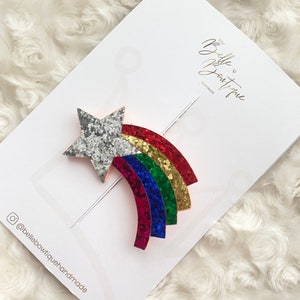Może przedstawiać: Gwiazda z błyszczącego tęczowego materiału z błyszczącą srebrną gwiazdą na g&oacute;rze. Tęcza wykonana jest z czerwonego, pomarańczowego, ż&oacute;łtego, zielonego, niebieskiego i fioletowego brokatu. Gwiazda znajduje się na białym tle z napisem "Belle Boutique Handmade" złotymi literami.