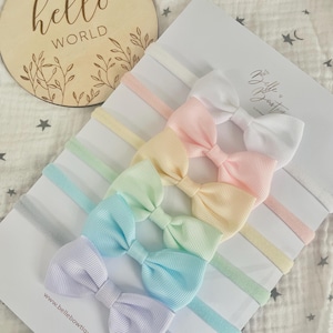 May include: Set of five pastel-coloured hair bows with matching elastic headbands. The bows are made of a soft, textured fabric and are arranged in a rainbow pattern. The headbands are made of a thin, stretchy material.