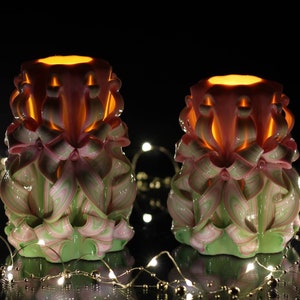 以下が含まれることがあります： 複雑な花柄のピンクとグリーンの装飾的なキャンドルが2本。キャンドルは灯され、輝いています。