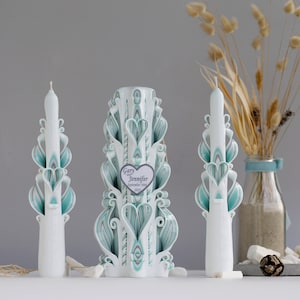 May include: A set of three white carved candles with teal and gray accents. The center candle has a heart-shaped tag with the text "Gary and Jennifer September 10th, 2023".