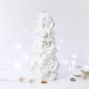 Puede incluir: Una vela blanca tallada con diseños florales y de cinta, y dos anillos dorados. La vela está decorada con rosas y corazones. El fondo es blanco con luces de hadas y elementos decorativos.