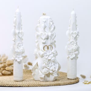 Puede incluir: Tres velas de boda blancas con intrincados diseños florales y de corazón. La vela del centro tiene dos anillos dorados colocados en la parte superior.