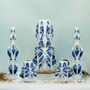 May include: A set of four white and blue wedding candles with intricate designs. The candles are decorated with gold rings and white pearls. The set includes a tall center candle and three smaller candles.
