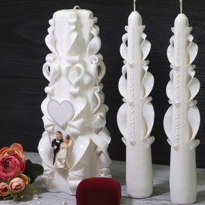 Könnte beinhalten: Drei weiße Hochzeitskerzen mit aufwendigen Schnitzereien. Die mittlere Kerze zeigt eine Paarskulptur und ein Herz. Die Kerzen stehen auf einer weißen Oberfläche mit einer roten Ringbox.