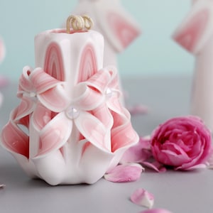 May include: A carved white candle with pink ribbon-like accents and clear crystal embellishments. The candle has a spiral design and is surrounded by a pink rose and petals. The candle has a twisted, beige wick.
