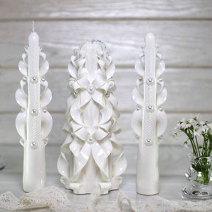 May include: A set of three white carved candles with pearl accents. The center candle is taller and features a more elaborate design with a floral motif. The candles are arranged on a white lace tablecloth.