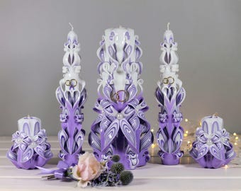 May include: A set of five white and purple carved candles for a wedding ceremony. The tallest candle is decorated with two gold rings and the smaller candles are decorated with pearls.