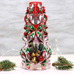 May include: A hand-carved Christmas candle with red, white, and green swirls. The candle is decorated with a pine cone, holly, and a gold bell.