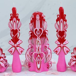 Puede incluir: Conjunto de cinco velas de boda rojas y blancas con diseños de corazones y cintas. La vela central es más alta y tiene un porta anillos.