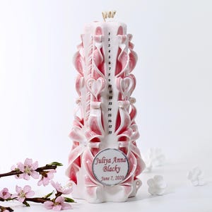 May include: Pink and white carved candle with a personalized label that reads "Juliya Anna Blacky June 7, 2020". The candle has numbers 1-18 carved on the side.