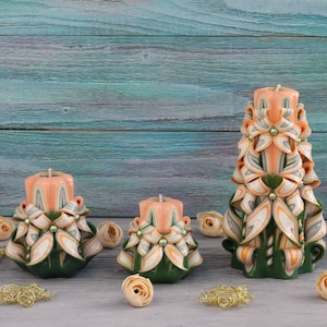 以下が含まれることがあります： 複雑な花柄が施された手彫りのキャンドルが3本。キャンドルはピーチ色のワックスでできており、緑の葉と白いアクセントが付いています。キャンドルは薄い灰色の表面に並べてあります。
