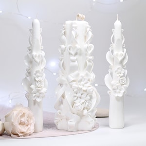 以下が含まれることがあります： 花とリボンのデザインが施された、白い彫刻キャンドルが3つ。キャンドルは高さが異なり、バラやハートの形など、複雑なディテールが特徴です。薄いピンク色のバラがいくつか前景にあります。