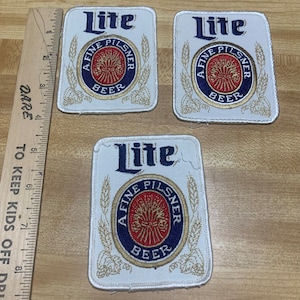 May include: Three white fabric patches with the words "Lite" and "A Fine Pilsner Beer" in gold and blue lettering. The patches are embroidered with a gold wheat design.