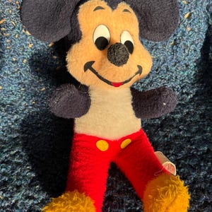 Op de afbeelding: Vintage pluche Mickey Mouse speelgoed met zwarte oren, een geel en wit gezicht, een rode broek met gele knopen en gele vacht op de voeten.