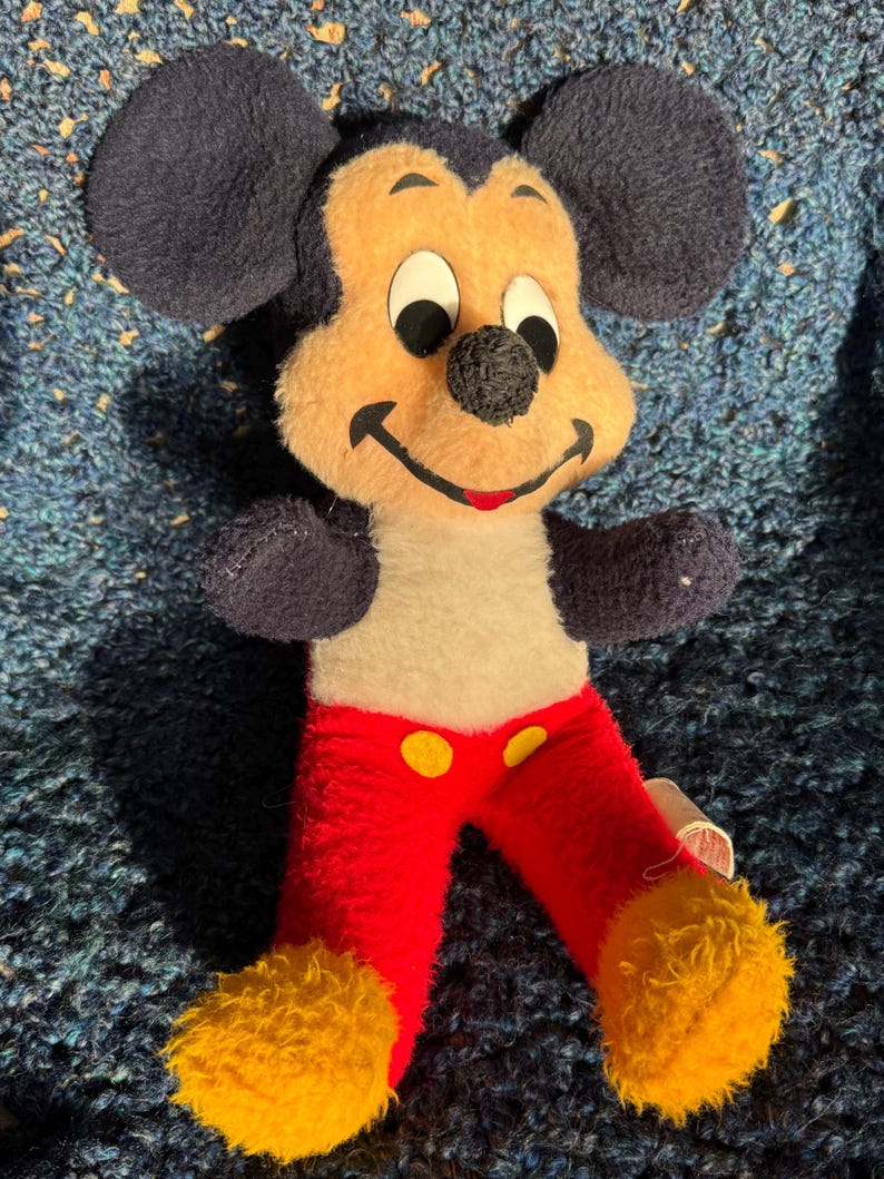 Op de afbeelding: Vintage pluche Mickey Mouse speelgoed met zwarte oren, een geel en wit gezicht, een rode broek met gele knopen en gele vacht op de voeten.