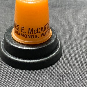 May include: Orange plastic thimble with the text "James E. McCarthy Diamonds, Watches" on the side. The thimble is sitting on a black plastic base.