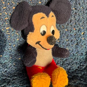 Op de afbeelding: Een pluche Mickey Mouse speelgoed dat op een blauw gehaakt oppervlak zit. Het speelgoed draagt een rode broek met gele schoenen en heeft zwarte oren en een zwarte neus.