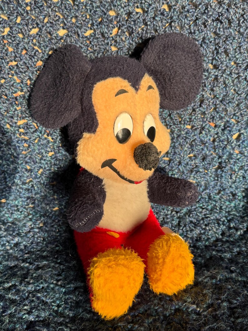 Op de afbeelding: Een pluche Mickey Mouse speelgoed dat op een blauw gehaakt oppervlak zit. Het speelgoed draagt een rode broek met gele schoenen en heeft zwarte oren en een zwarte neus.
