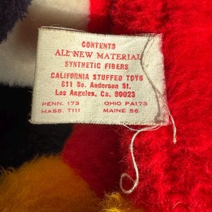 Op de afbeelding: Een wit label met zwarte tekst die "CONTENTS ALL NEW MATERIAL SYNTHETIC FIBERS CALIFORNIA STUFFED TOYS 611 So. Anderson St. Los Angeles, Ca. 90023 PENN. 173 MASS. T111 OHIO PA173 MAINE 56" luidt.