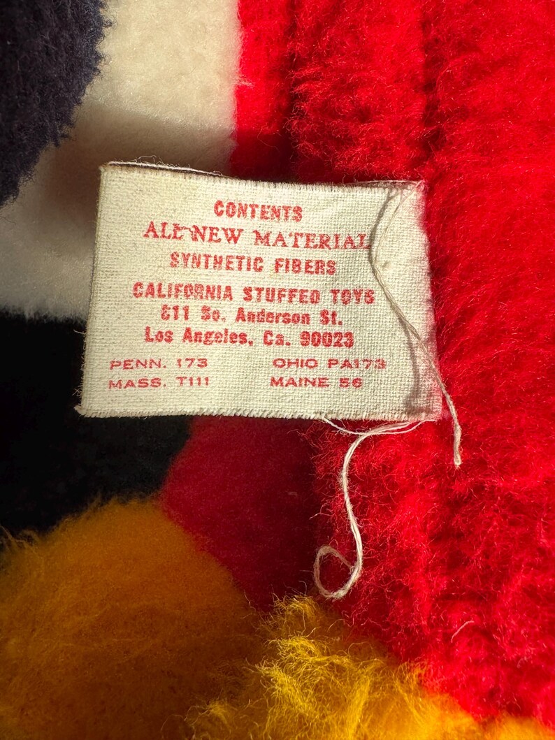 Op de afbeelding: Een wit label met zwarte tekst die "CONTENTS ALL NEW MATERIAL SYNTHETIC FIBERS CALIFORNIA STUFFED TOYS 611 So. Anderson St. Los Angeles, Ca. 90023 PENN. 173 MASS. T111 OHIO PA173 MAINE 56" luidt.