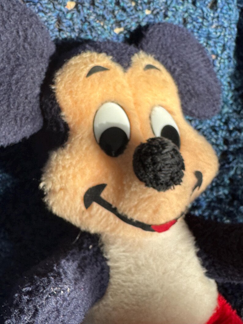 Op de afbeelding: Een pluche Mickey Mouse speelgoed met zwarte oren, een zwarte neus en witte ogen. Het speelgoed draagt een rood-witte outfit.