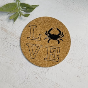 以下が含まれることがあります： 「LOVE」の文字と黒いカニのシルエットが入った丸いコルクコースター。コースターは自然なタン色で、白い木の表面に置かれています。緑の葉の小枝が左上にあります。