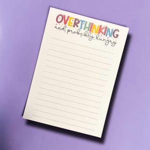 May include: A lined notepad with the words "OVERTHINKING and probably hungry" in colorful lettering. The notepad is white with horizontal gray lines and sits on a purple background. The text is in a playful, handwritten style.