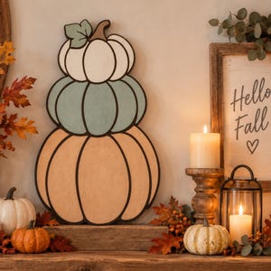 May include: A decorative wall hanging featuring three stacked pumpkins in white, sage green, and light orange with black outlines. The text "Fall Stacked Pumpkins" and "Finished size is 18.5" H x 10.75" W" is printed at the bottom of the image.